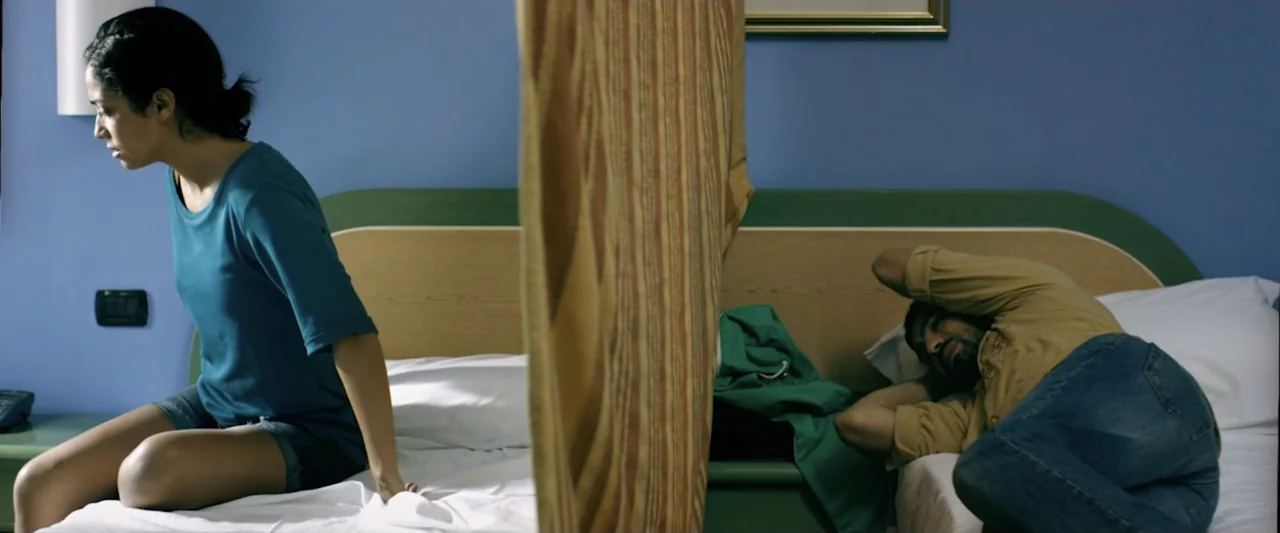Taranta On the Road (2017) movie still: medium — A hotel room with blue walls features two separate beds divided by a curtain. A woman sits on one…