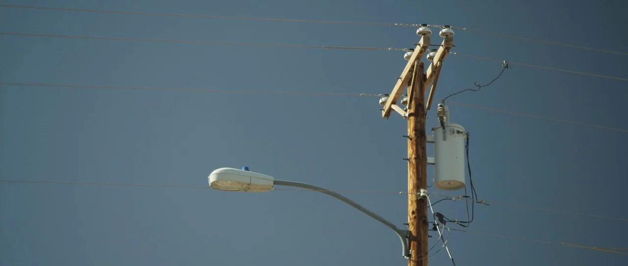 Frank (2014) movie still: wide — A wooden utility pole stands against a clear, solid blue sky. Several power lines extend horizontally across the…