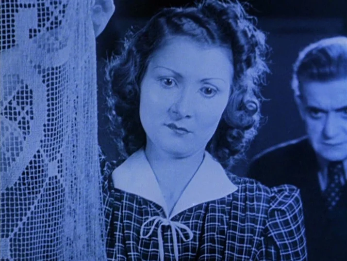 Innocence Unprotected (1968) movie still: medium — A woman stands in the foreground holding a delicate white lace curtain. An out-of-focus man in a…