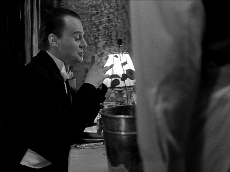 My 20th Century (1989) movie still: medium — A man in a formal tuxedo sits at a dining table at night. He gestures with his hands while looking…
