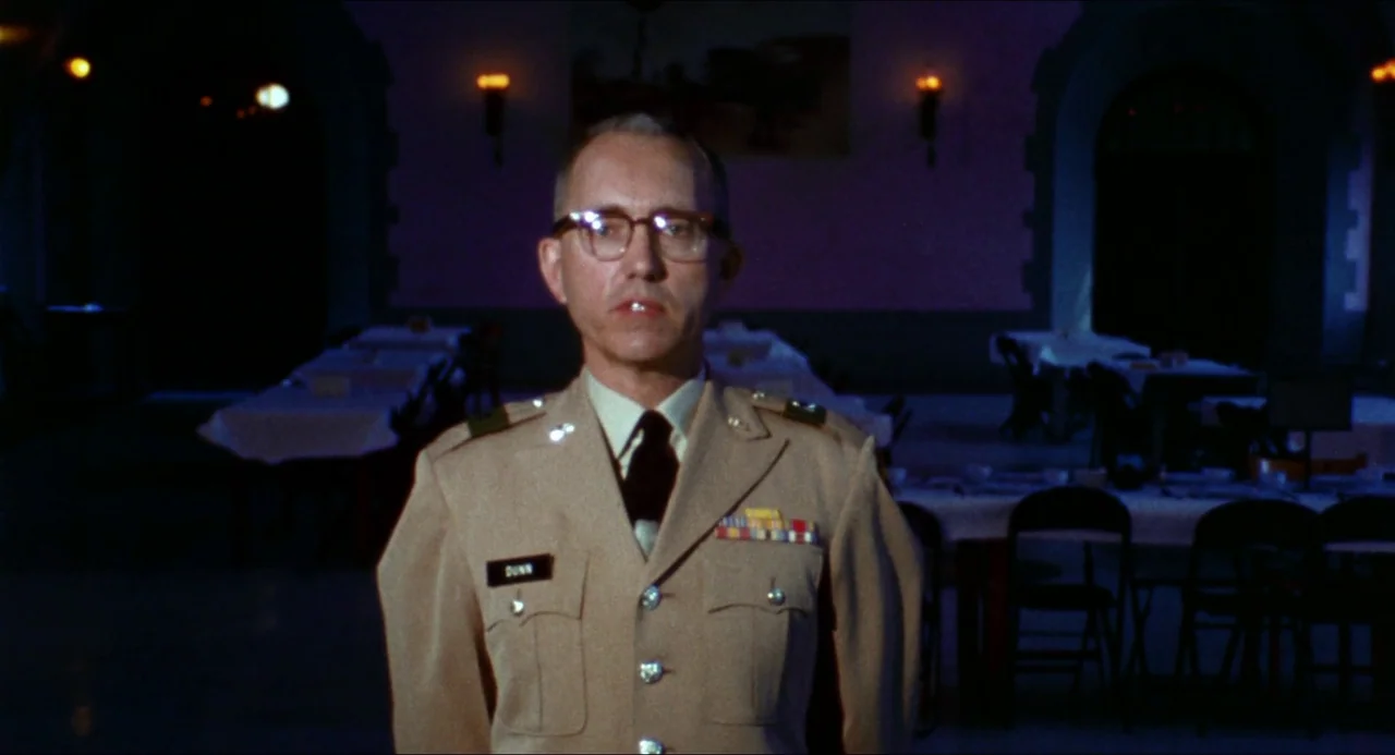 Medium Cool (1969) movie still: medium — A dark, empty mess hall with rows of tables draped in white cloths. A man in military uniform stands…