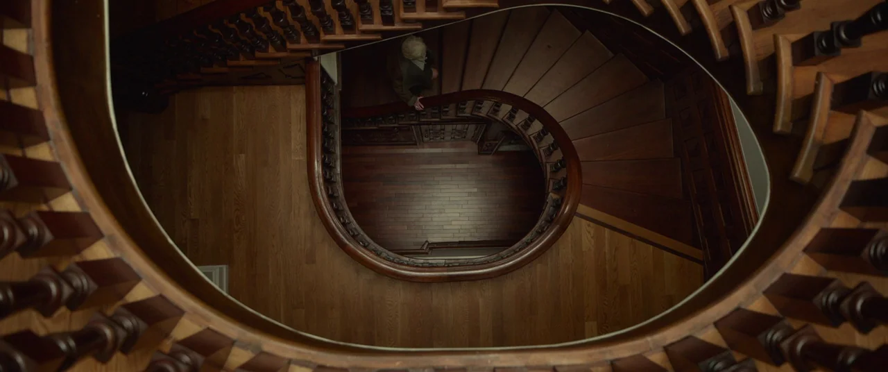 Split (2016) movie still: wide — A curving wooden staircase with ornate balusters spirals downward into an open foyer. A woman stands on the…
