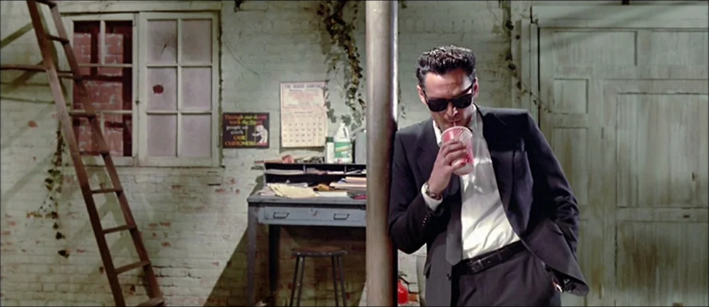 Reservoir Dogs (1992) movie still: medium — An industrial space features painted white brick walls and a metal desk with papers. A man in a dark…