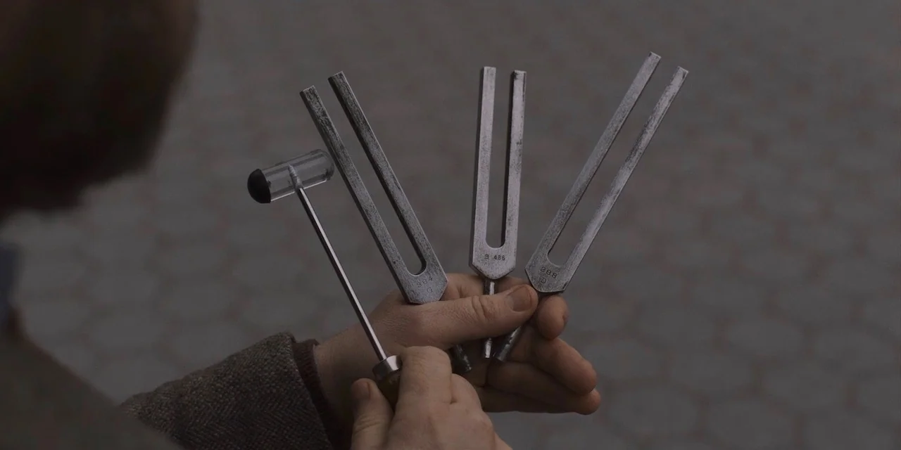 The Sound of Silence movie still: closeup — A person holds three metal tuning forks and a transparent plastic reflex hammer. The objects are…