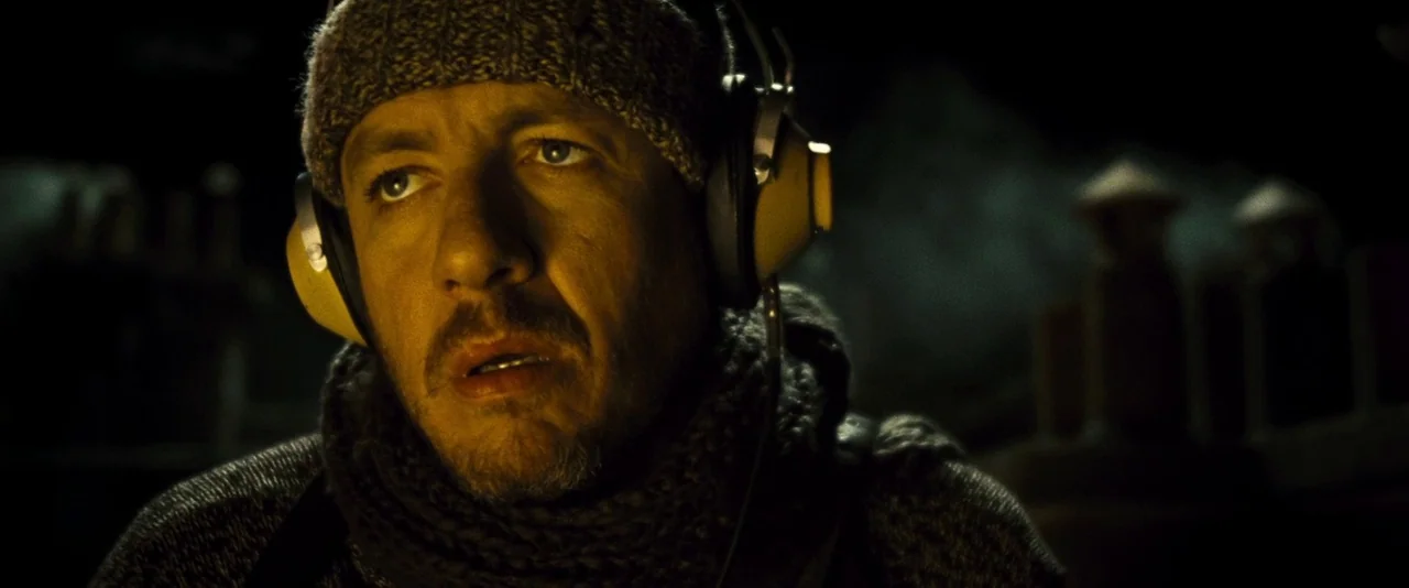 Micmacs (2009) movie still: closeup — A man wearing winter headwear and headphones occupies a dark, undefined interior space. He looks toward the…