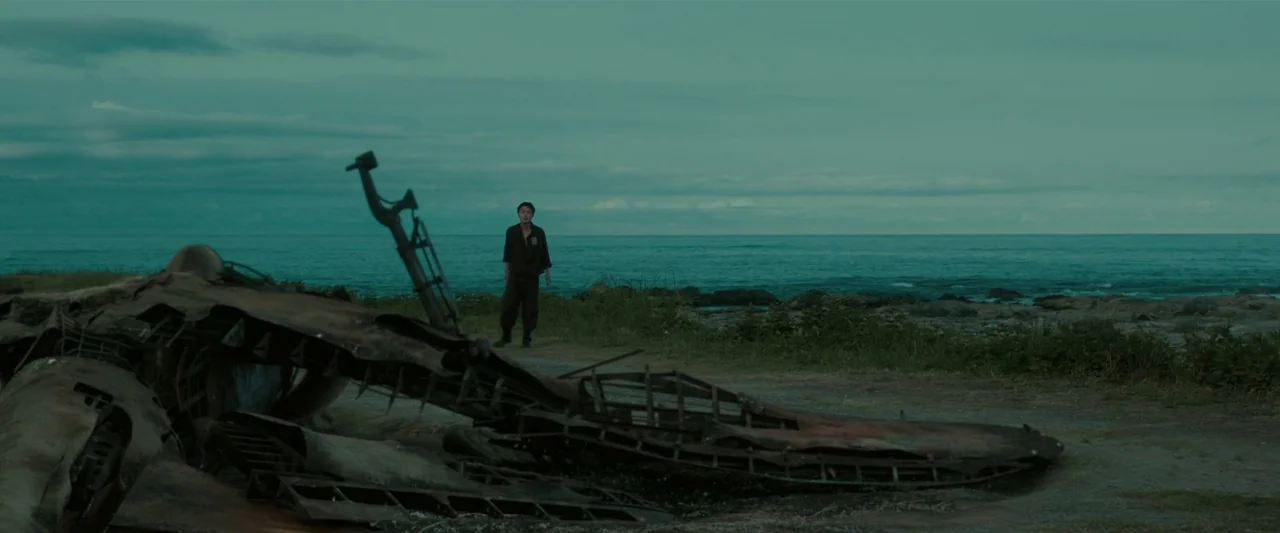 Godzilla Minus One (2023) movie still: wide — A desolate rocky coast features a wrecked airplane fuselage resting on the ground. A young man stands…