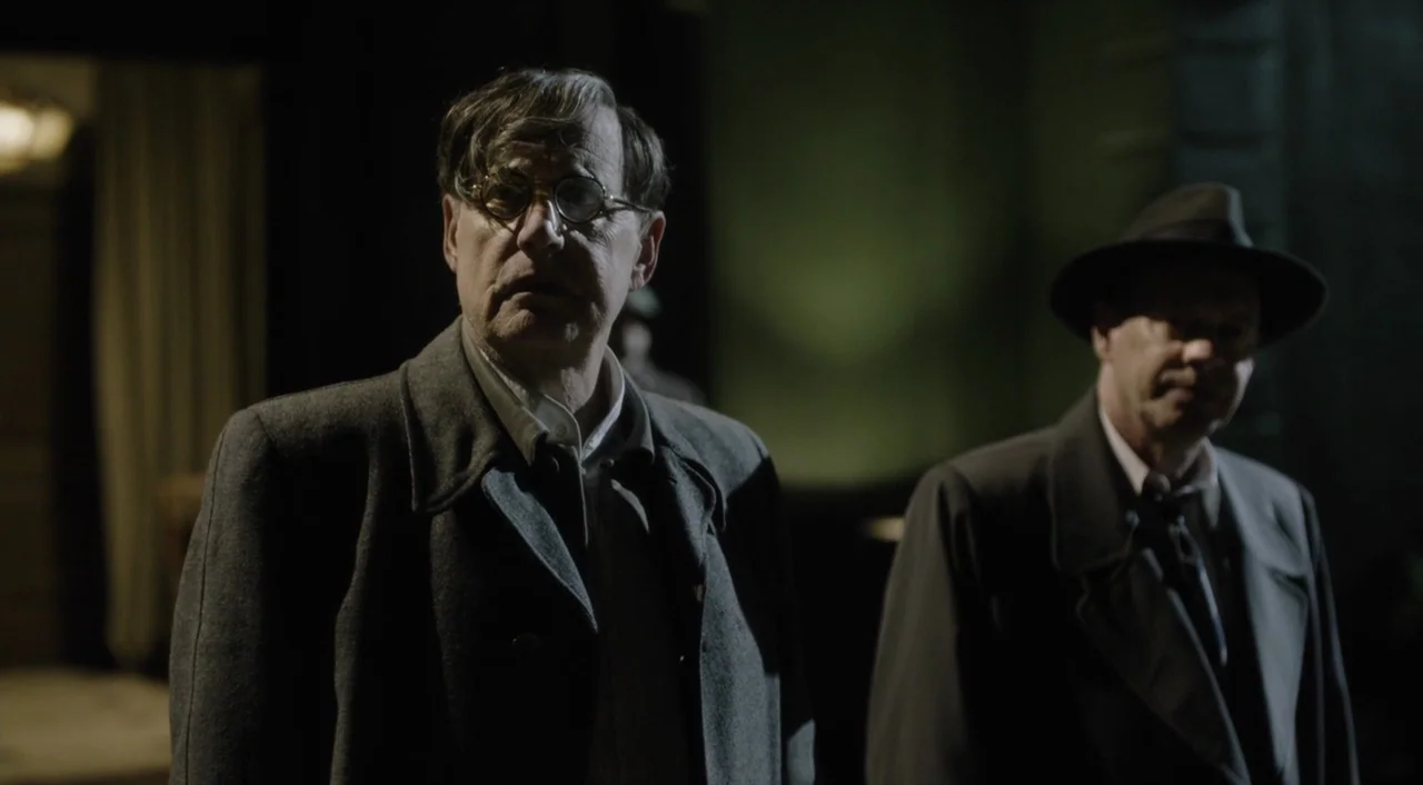 The Death of Stalin (2017) movie still: medium — A dark, shadowed corridor serves as the backdrop for two men in coats. One man in glasses stands in…