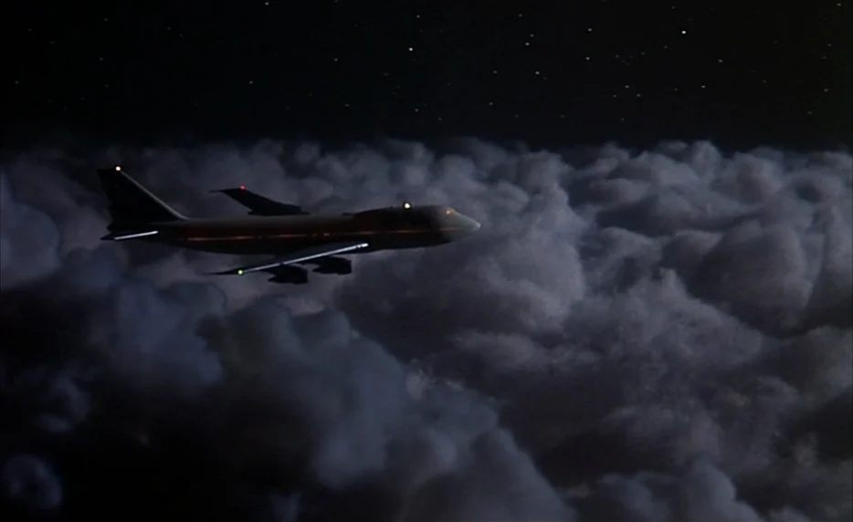 I Want To Go Home (1989) movie still: wide — A dark night sky over an expansive, thick bank of rolling clouds. A passenger jet travels horizontally…