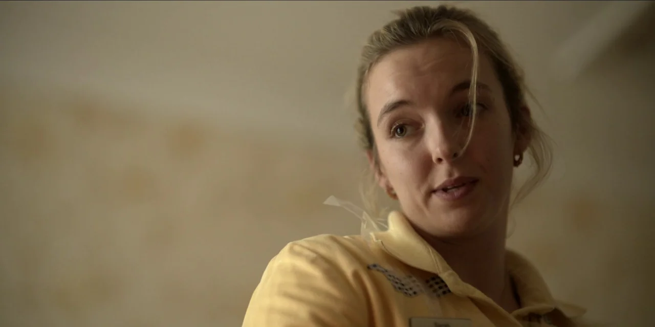 Help (2021) movie still: closeup — A woman stands in a nondescript indoor space with light-colored walls. She wears a yellow work uniform while…