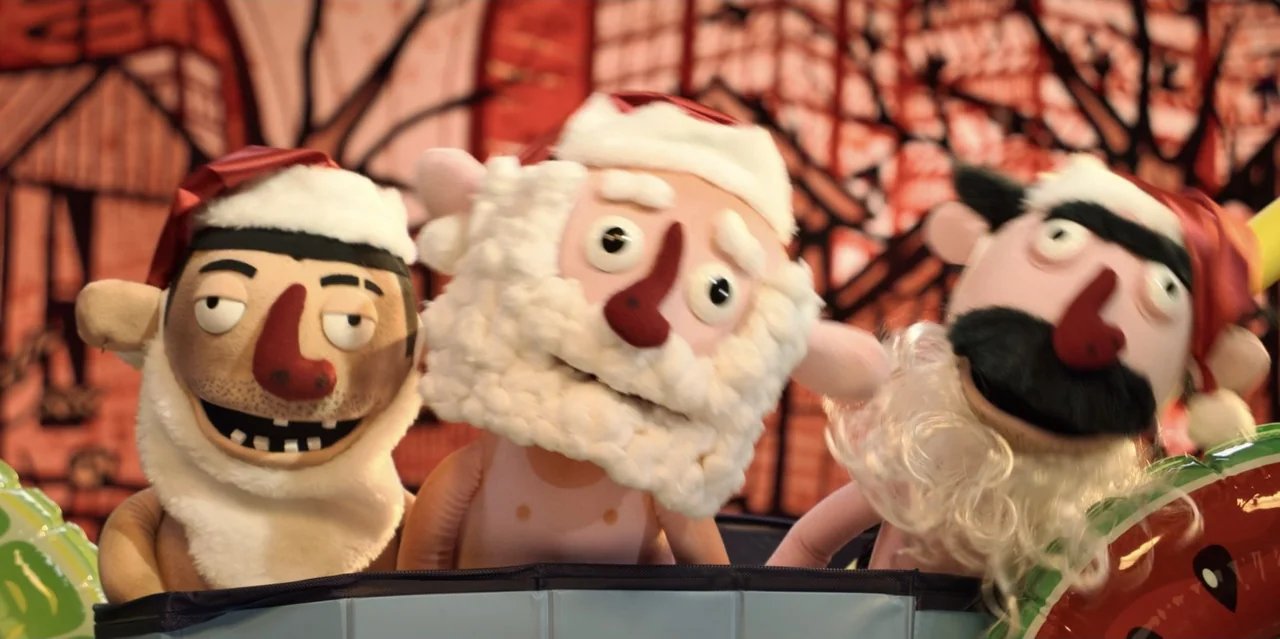 31 Minutes: One Hot Christmas (2025) movie still: medium — Three puppets wearing Santa hats stand against a patterned red and white backdrop. The…