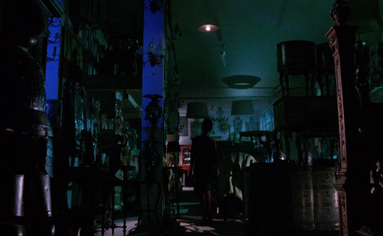 Blood And Black Lace (1964) movie still: wide — A dark antique shop is filled with cluttered furniture pieces under deep blue lighting. A person…
