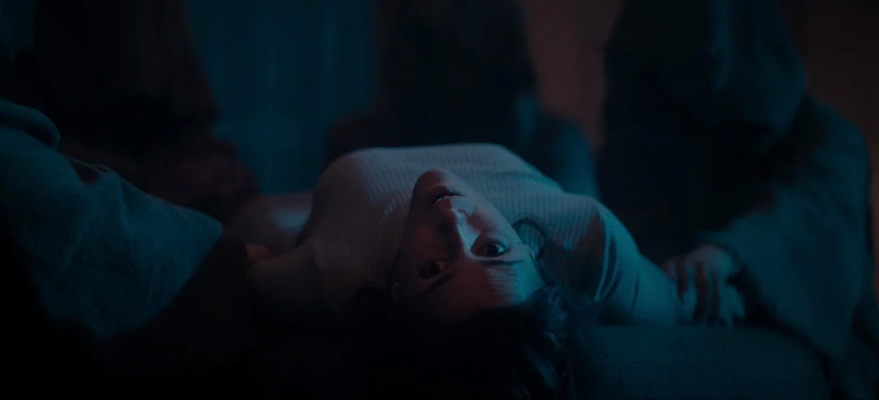 Housewife (2017) movie still: medium — A woman lies on her back in a dark room surrounded by hooded figures. The scene is illuminated by cold blue…