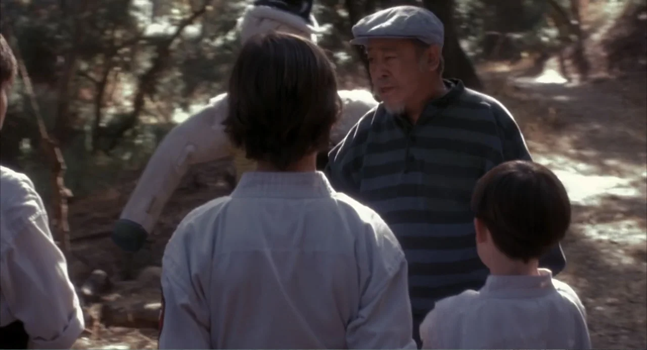 3 Ninjas (1992) movie still: medium — A group stands on a dirt path in a wooded area with trees surrounding them. The man faces two children in…