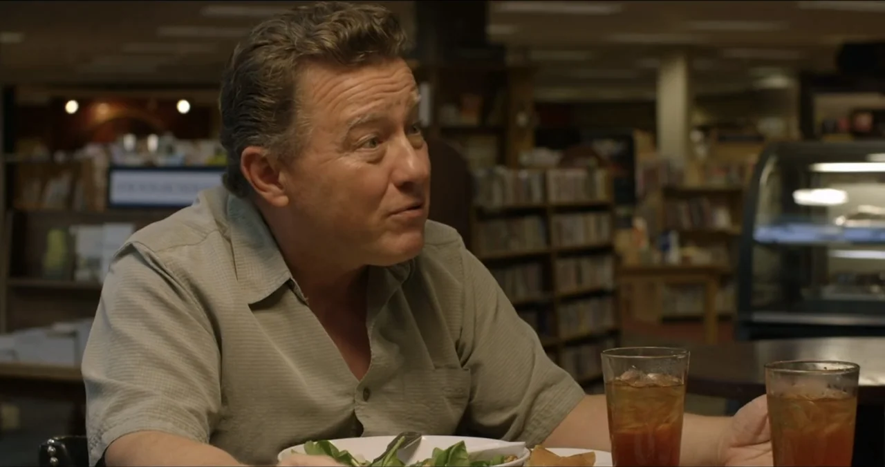Faith, Love & Chocolate (2018) movie still: medium — A man sits at a table inside a dimly lit restaurant with bookshelves. He is speaking while…