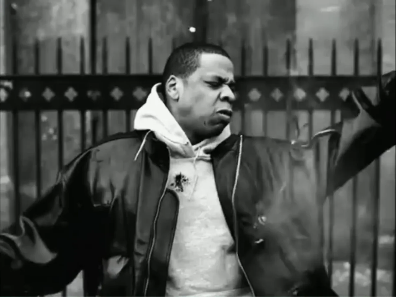 As We Speak: Rap Music on Trial (2024) movie still: medium — A man stands before a tall dark metal fence in an urban setting. He gestures outward…