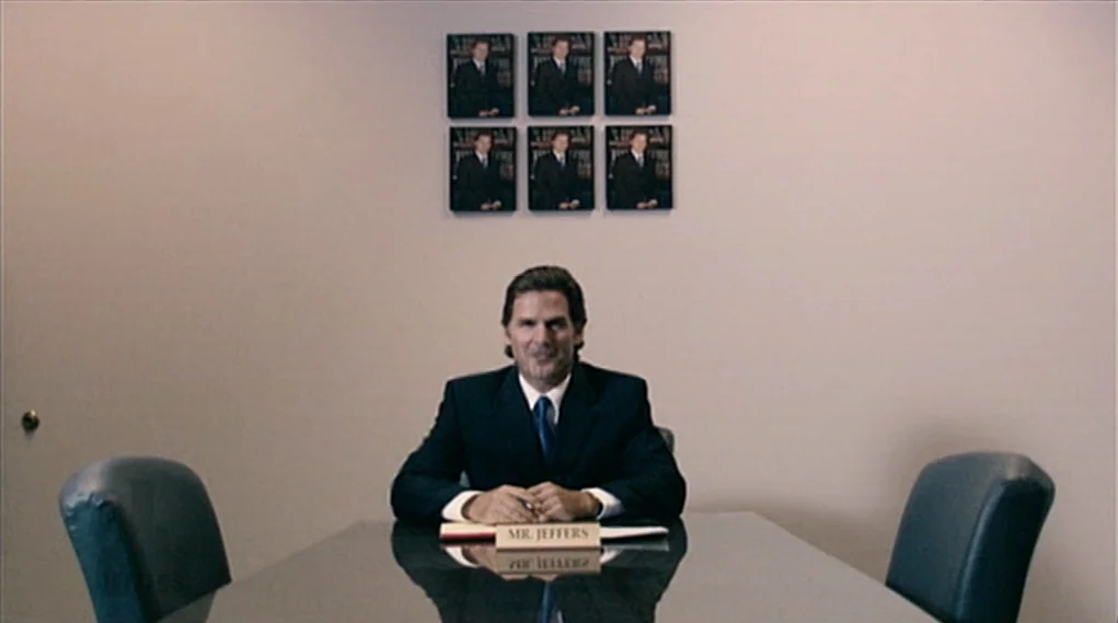 Visioneers (2008) movie still: medium — A man sits alone at a glossy table in a plain, beige-walled room. Six identical small framed photographs are…