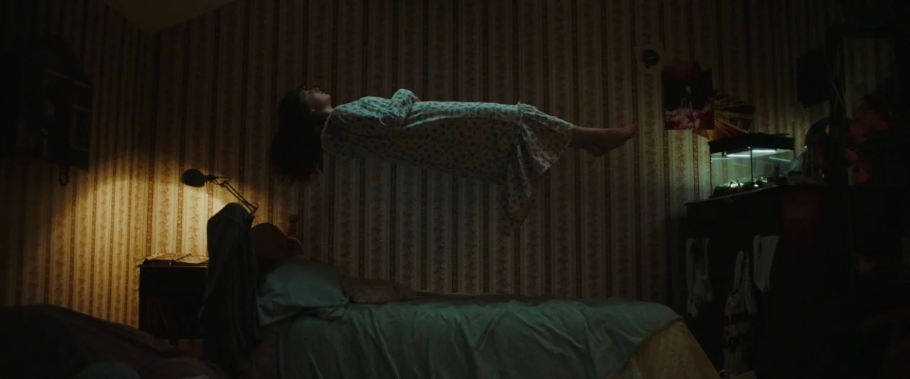 Extra Ordinary (2019) movie still: medium — A dimly lit room with striped wallpaper features a bed centered in the frame. A girl floats horizontally…