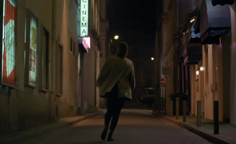 I Can’t Sleep (1994) movie still: fullbody — A narrow dark urban passage at night features a neon sign reading cinema. A woman in a blazer jogs away…