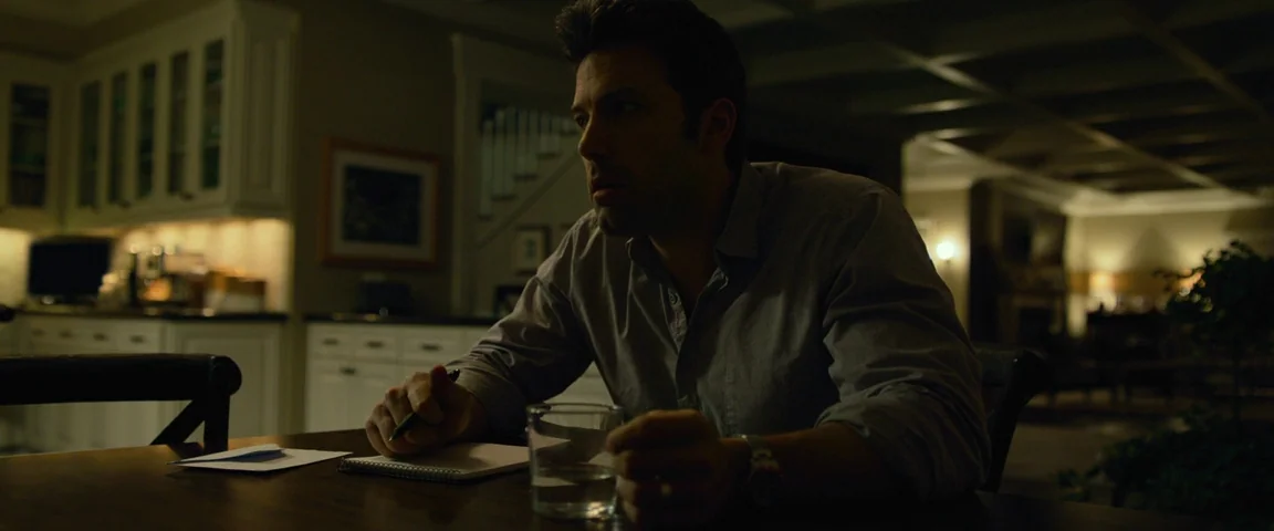Gone Girl (2014) movie still: medium — A dimly lit dining area with dark wooden furniture and white cabinetry in the back. A man sits alone at the…