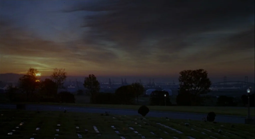 The Crossing Guard (1995) movie still: establishing — A sprawling cemetery with rows of grave markers stretches toward an industrial harbor horizon.…