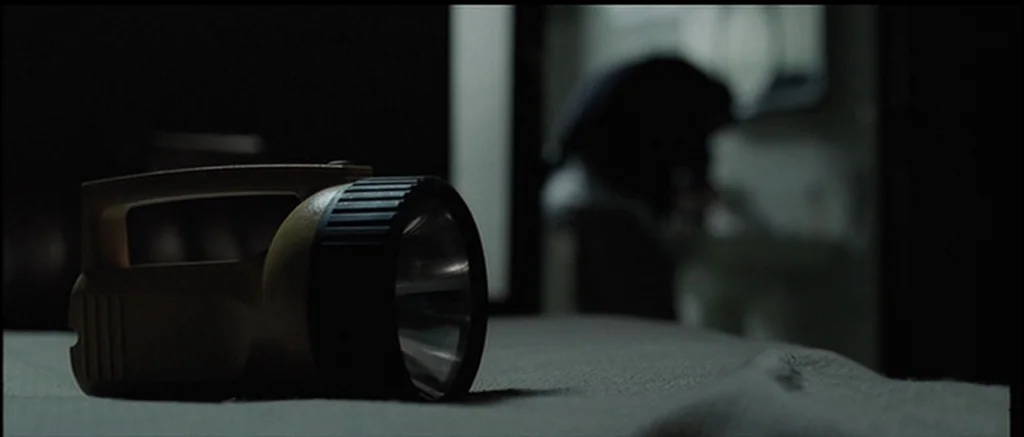 The Machinist (2004) movie still: closeup — A dimly lit room with a yellow handheld flashlight resting on a textured surface. A person is visible as…