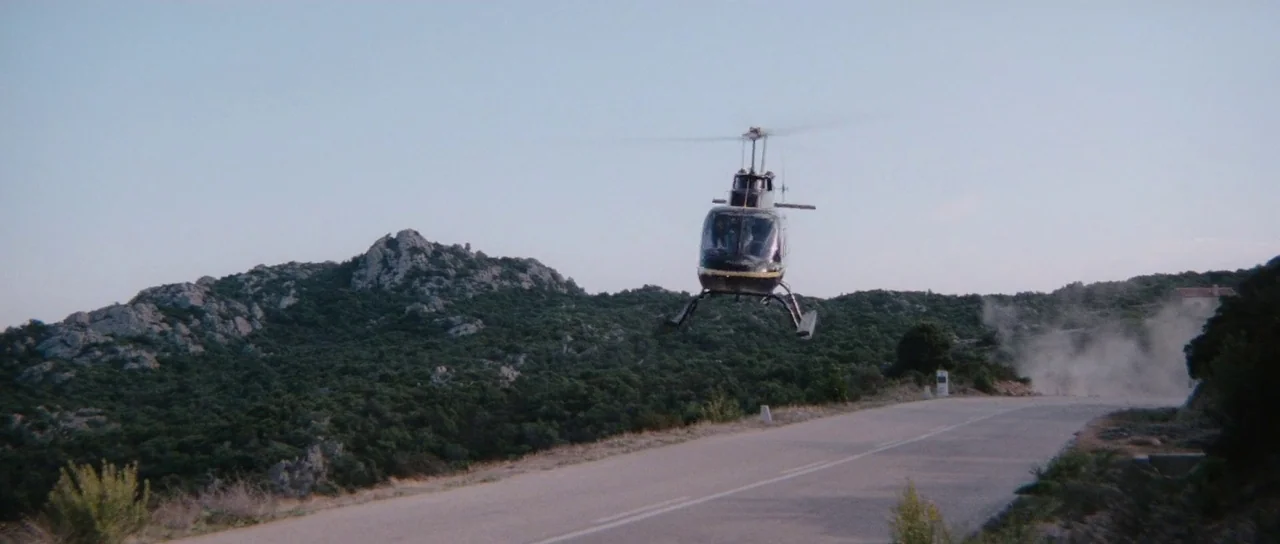 The Spy Who Loved Me (1977) movie still: wide — A dark helicopter maneuvers low over a rural two-lane road near hilly terrain. Dust rises from the…