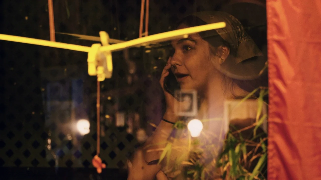 Gasoline Rainbow (2023) movie still: closeup — A person visible behind a glass partition at night with blurred foreground objects. The subject is…