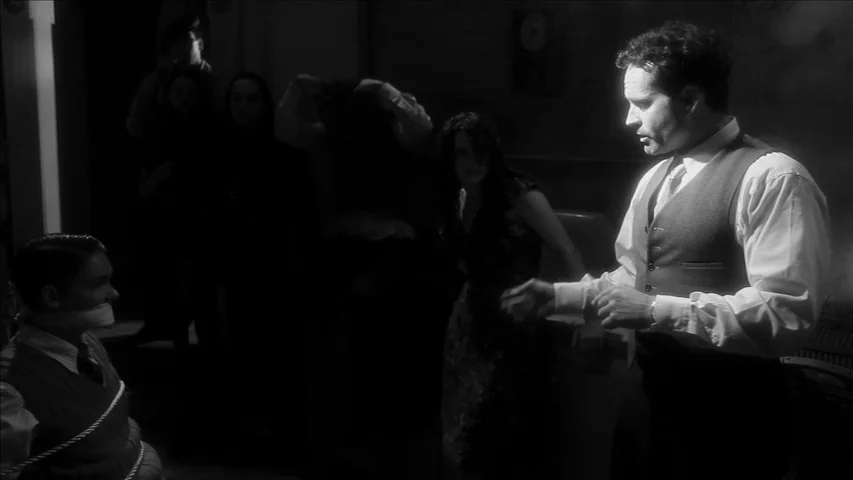 Keyhole (2011) movie still: medium — A dimly lit, shadowed basement setting filled with multiple indistinct figures in the background. In the…
