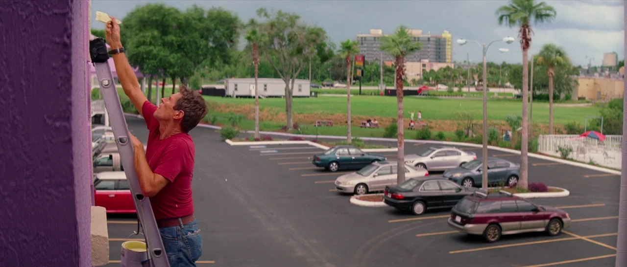 The Florida Project (2017) movie still: medium — A man stands on a metal ladder against a bright purple wall outdoors. A paved parking lot with…