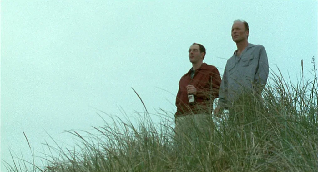 Pollock (2000) movie still: fullbody — A grassy sand dune under a completely overcast, pale sky. Two men stand side-by-side while looking into the…