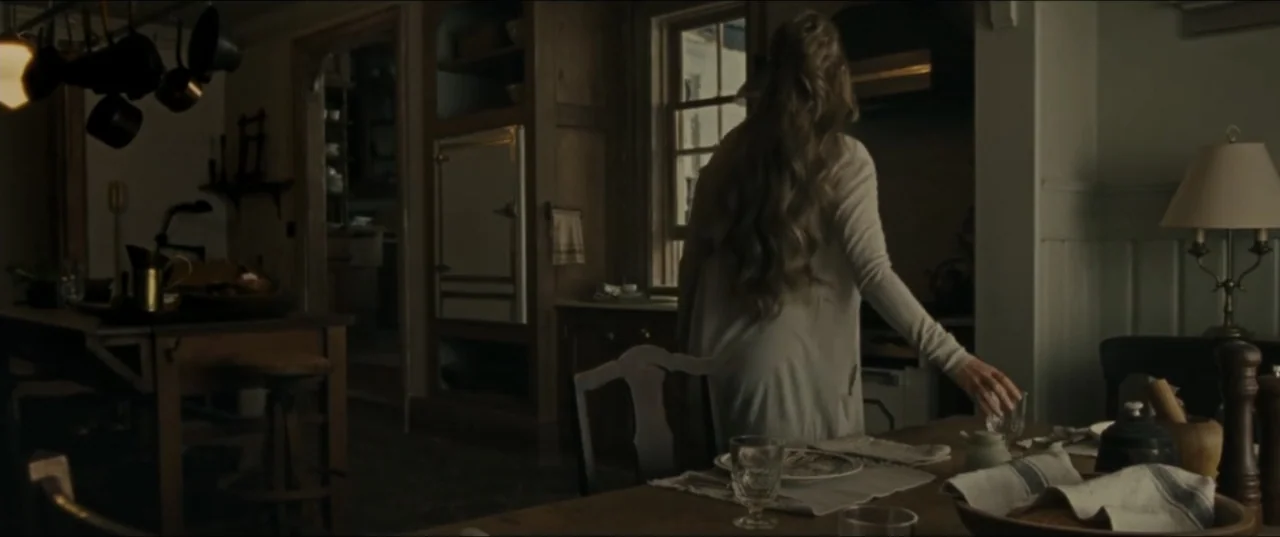 mother! (2017) movie still: medium — A dimly lit room features a wooden dining table and vintage architectural details. A woman in a light sweater…