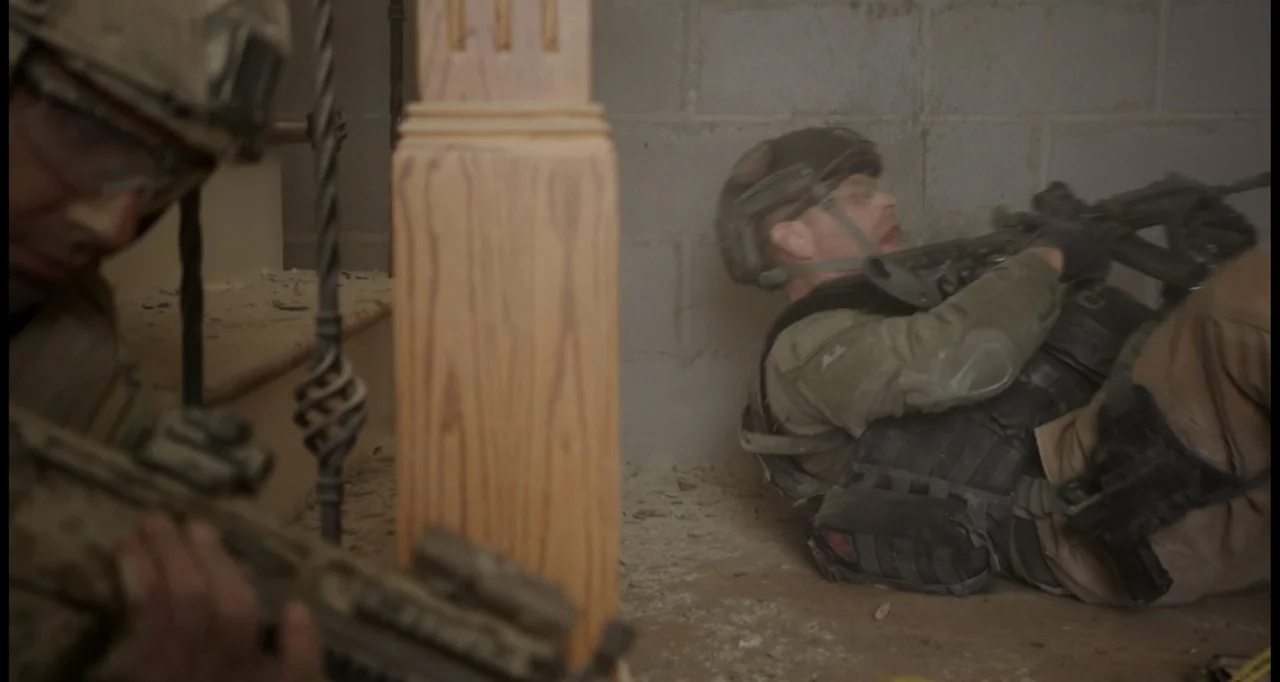 Sabotage (2014) movie still: medium — A soldier lies on concrete steps near a wooden baluster in a dusty room. Another soldier with a rifle stands…