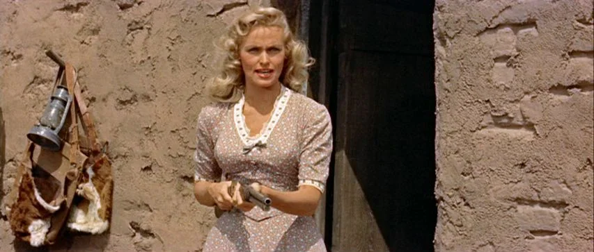Ride Lonesome (1959) movie still: medium — An adobe building wall with a rough texture serves as the background for a woman. The woman stands in an…