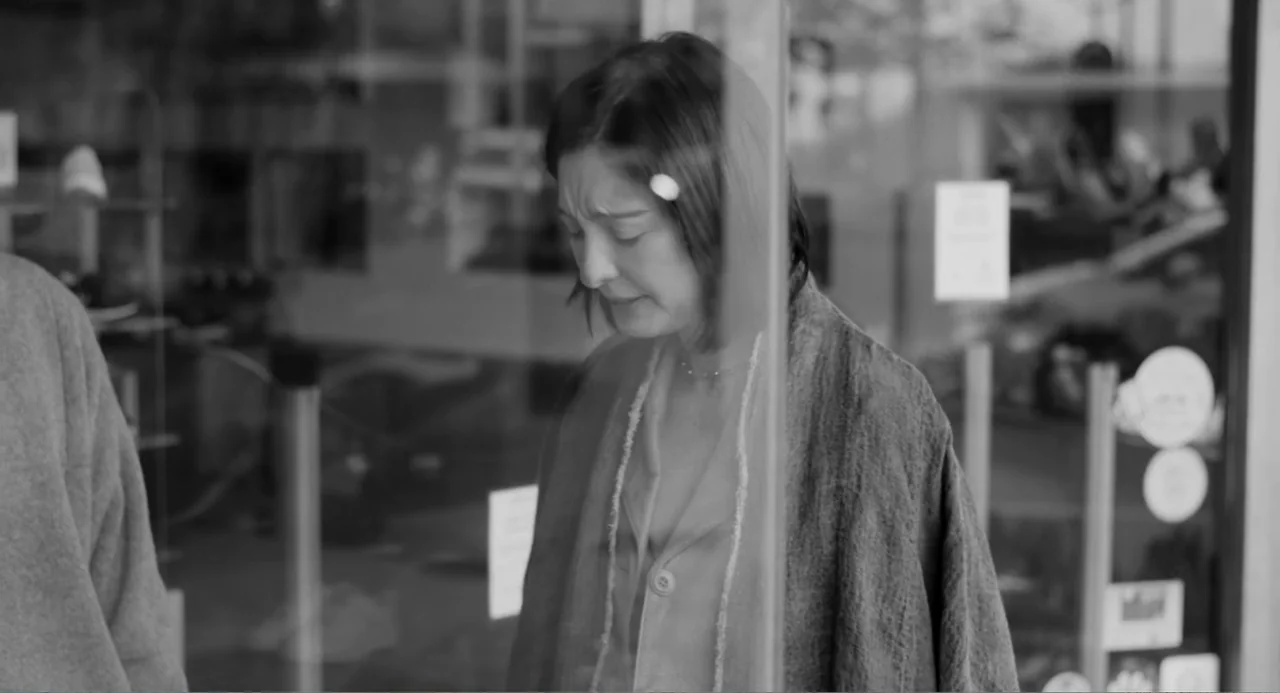 El Planeta (2021) movie still: medium — A woman stands behind a large glass storefront window during the day. The woman is positioned with her body…