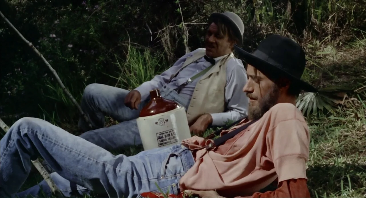 Cottonpickin' Chickenpickers (1967) movie still: medium — Two men recline on a grassy embankment in an outdoor area surrounded by thick vegetation.…