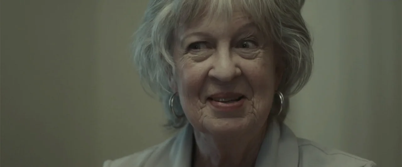 Jump, Darling (2022) movie still: closeup — A close-up view of an elderly woman in a sparsely decorated indoor room. The woman looks slightly away…