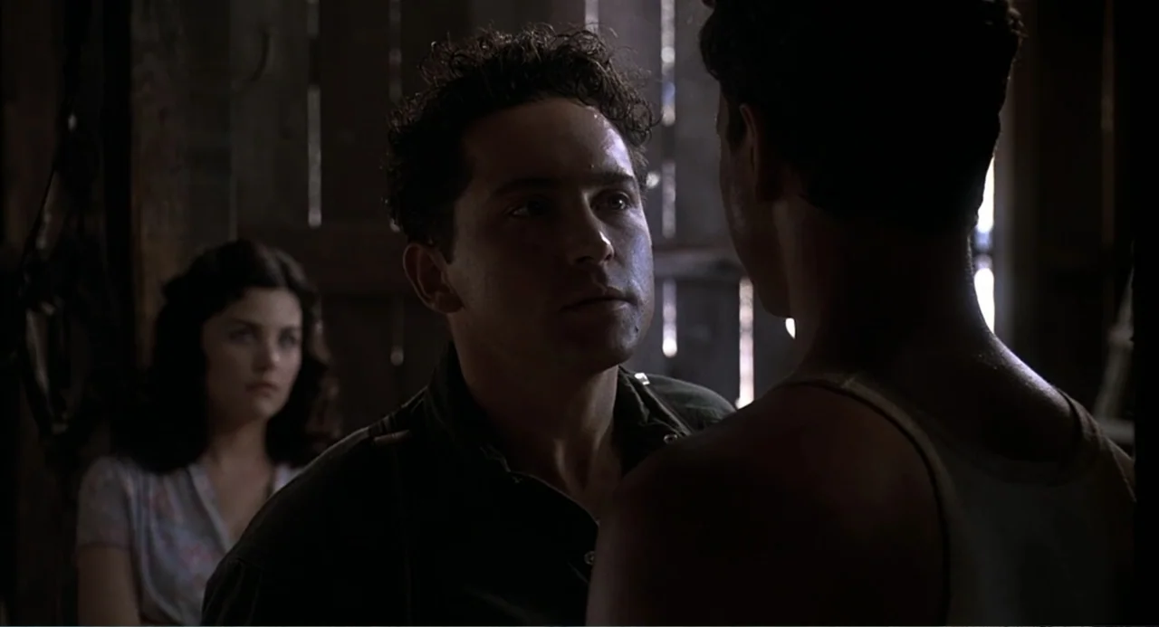 Of Mice and Men (1992) movie still: medium — The interior of a dimly lit barn features rough wooden plank walls and slats. A man stares at his…