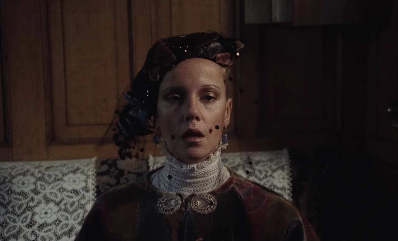 Mahler (1974) movie still: closeup — A woman sits against a wooden paneled wall with lace fabric behind her. She faces forward while wearing a…