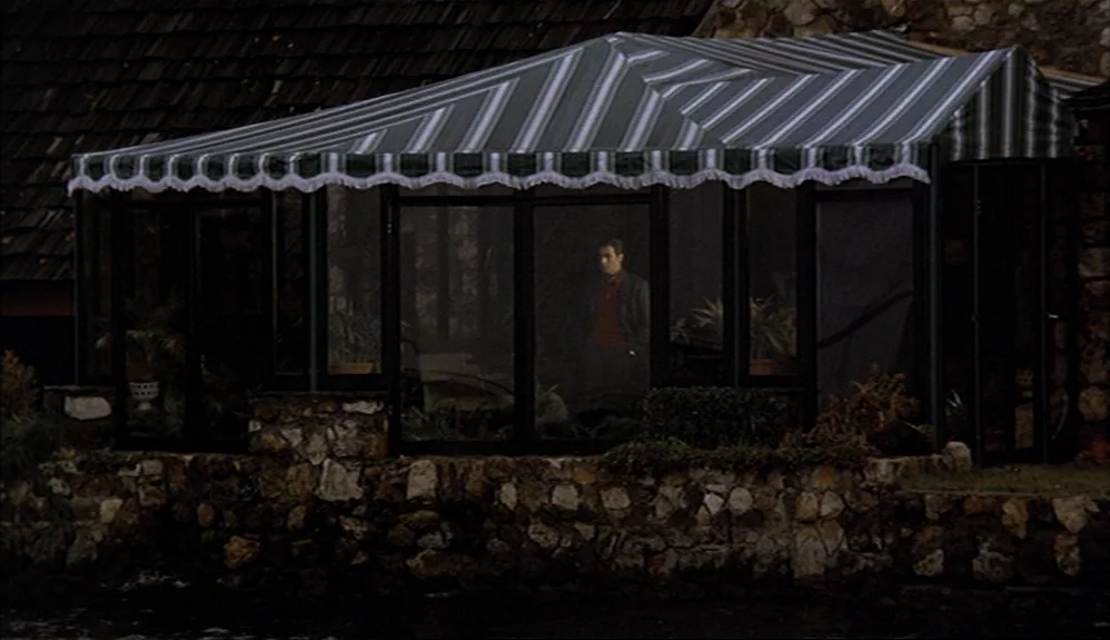 The Godfather Part II (1974) movie still: wide — A glass-walled sunroom with a green and white striped fabric awning sits by water. A man stands…