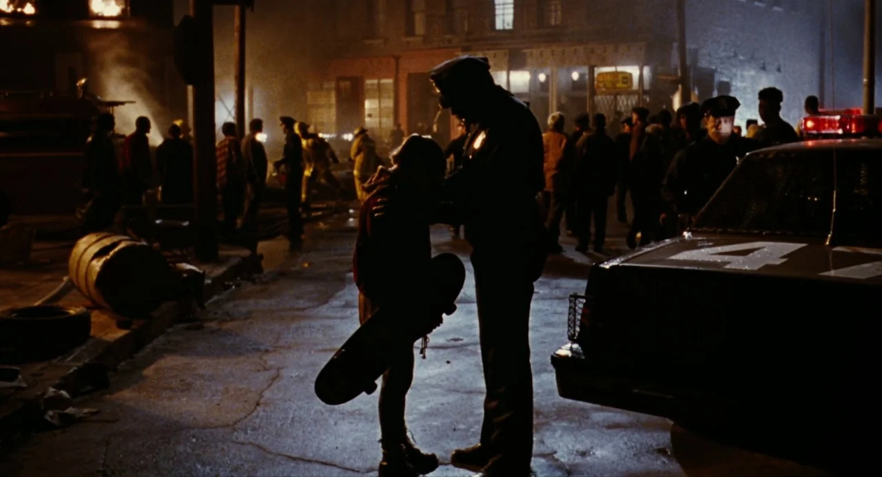 The Crow (1994) movie still: medium — A nighttime city street scene illuminated by the glow of a distant fire. A police officer stands beside a…