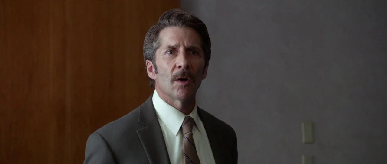 Faults (2014) movie still: medium — An indoor office with wood paneling on one side and a neutral gray wall. A middle-aged man with a mustache in a…