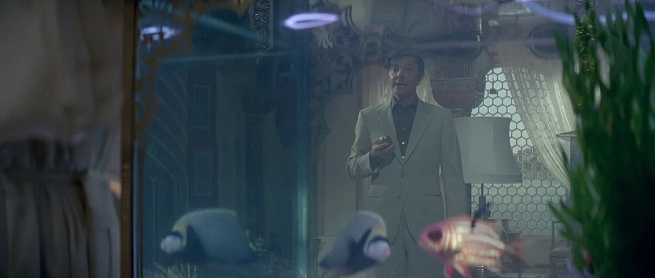Octopussy (1983) movie still: medium — A man stands in a room viewed through the glass of a fish tank. Fish swim in the foreground while the man…
