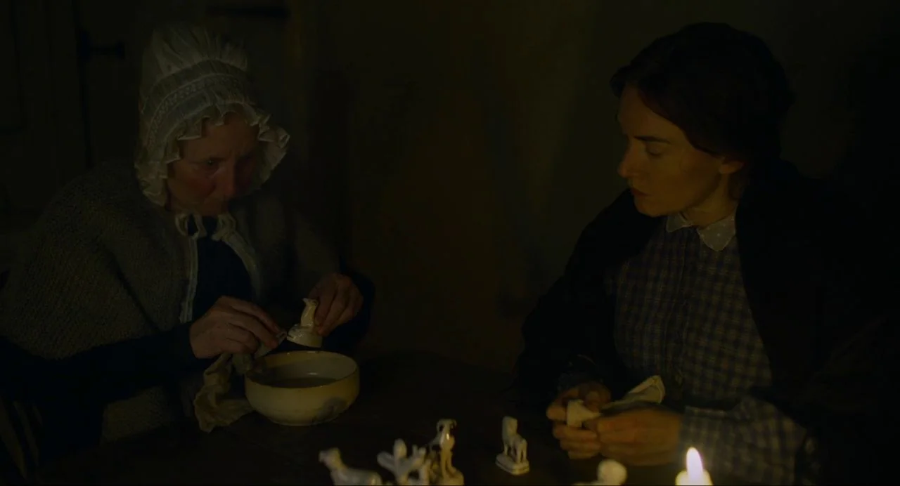 Ammonite (2020) movie still: medium — A dark, dimly lit room holds two women working together at a rustic table. The older woman cleans a small…