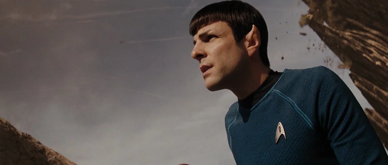Star Trek (2009) movie still: medium — A rocky, barren landscape under a dusty, pale sky with falling debris. A man with pointed ears wearing a blue…