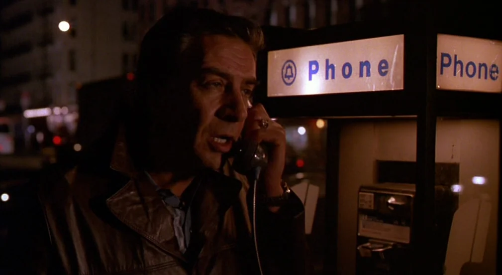 Crimes and Misdemeanors (1989) movie still: medium — An outdoor city sidewalk at night featuring a lit public payphone booth. A mature man in a…