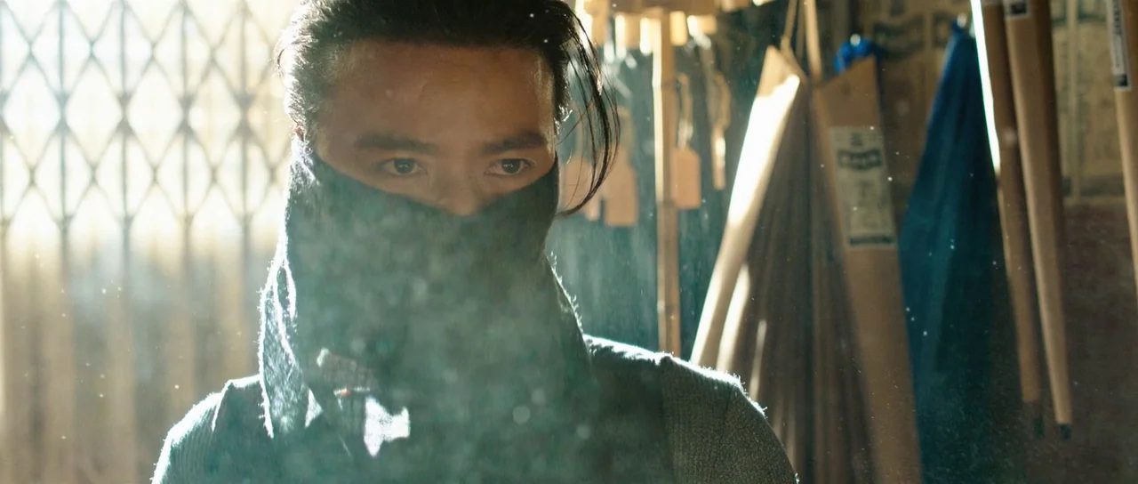 Ip Man 3 (2015) movie still: closeup — A cluttered workshop setting filled with wooden tools and hanging paper rolls. A young man wearing a dark…