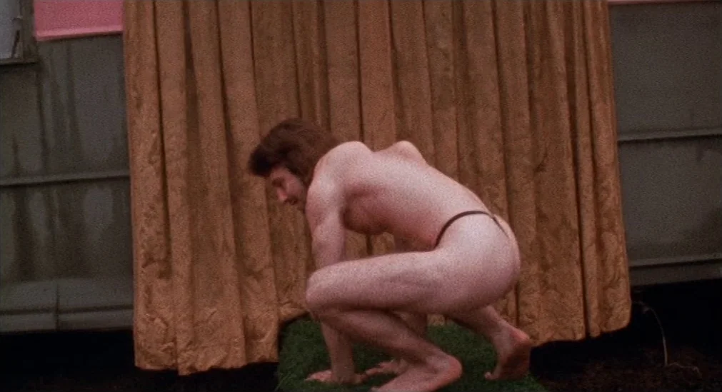 Pink Flamingos (1972) movie still: medium — A man crouches on a small patch of artificial turf against a golden curtain. He is mostly nude and…