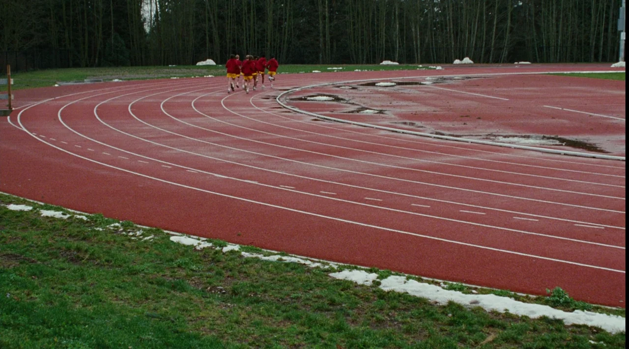 Juno (2007) movie still: wide — An outdoor red athletic running track features remnants of melting snow along the edges. A small group of runners…