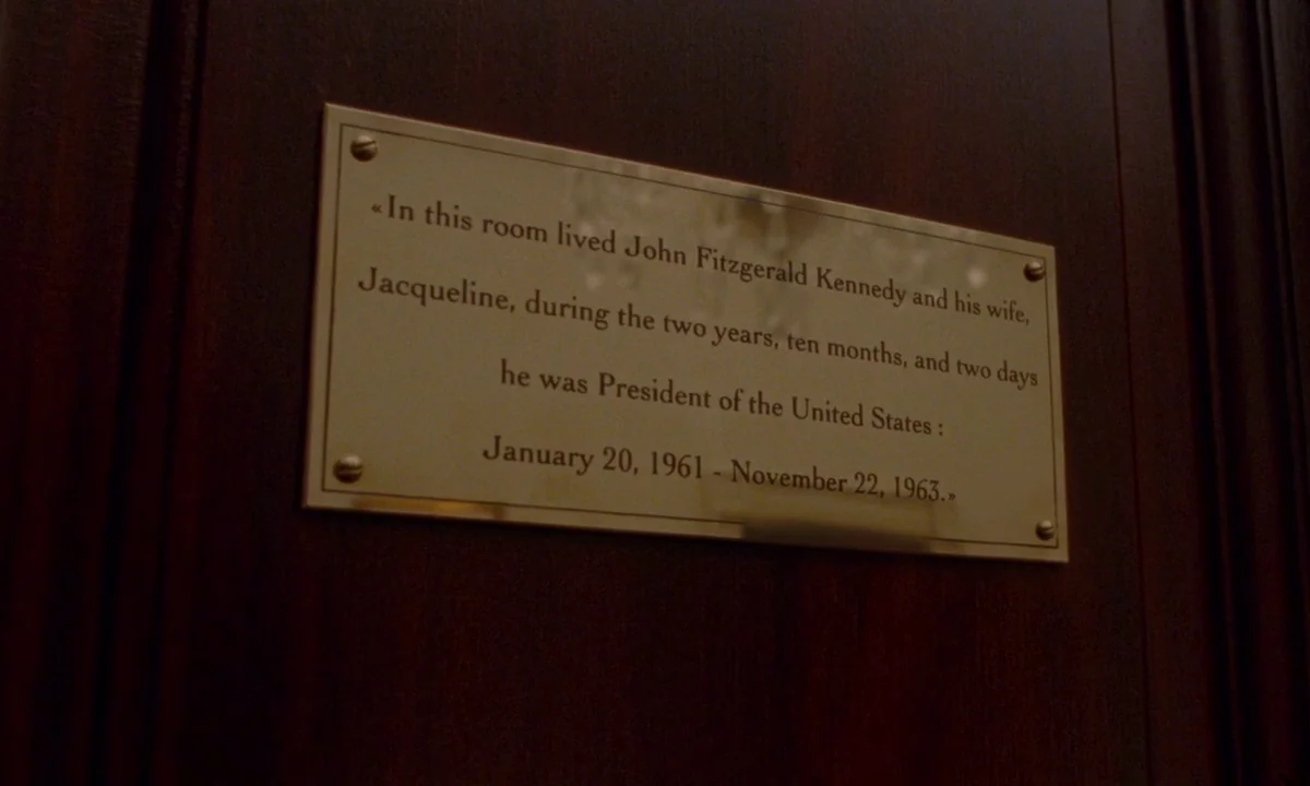 Jackie (2016) movie still: closeup — A brass memorial plaque is mounted onto a dark, polished wooden surface. The inscription recounts the residency…