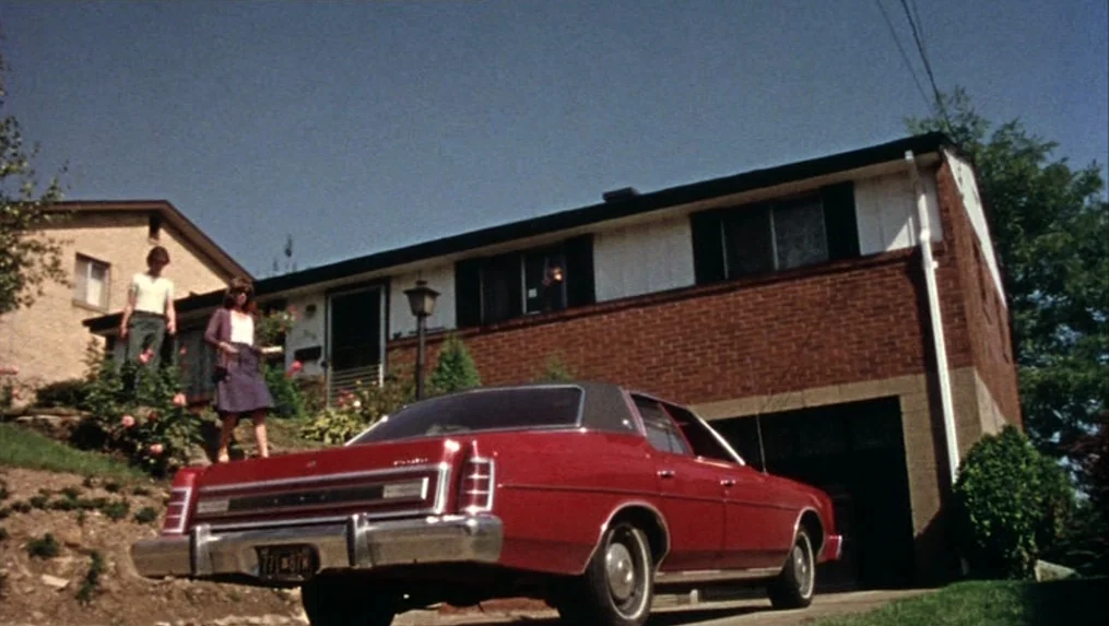 Martin (1977) movie still: wide — A split-level suburban home features a brick facade with a white upper story. A young man and woman walk down the…